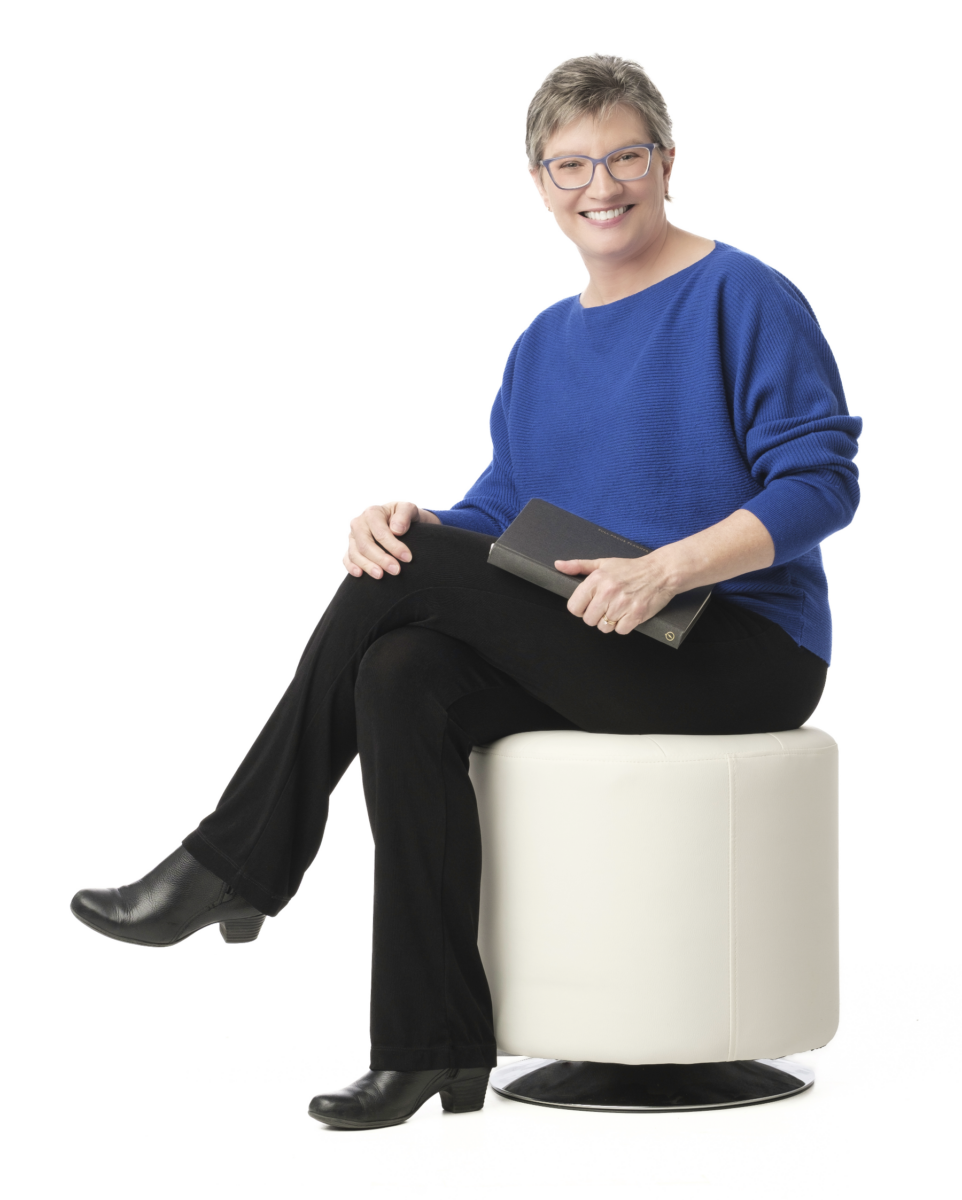 Kathy J. Lang sitting on white stool holding notebook and smiling, wearing blue sweater and black slacks